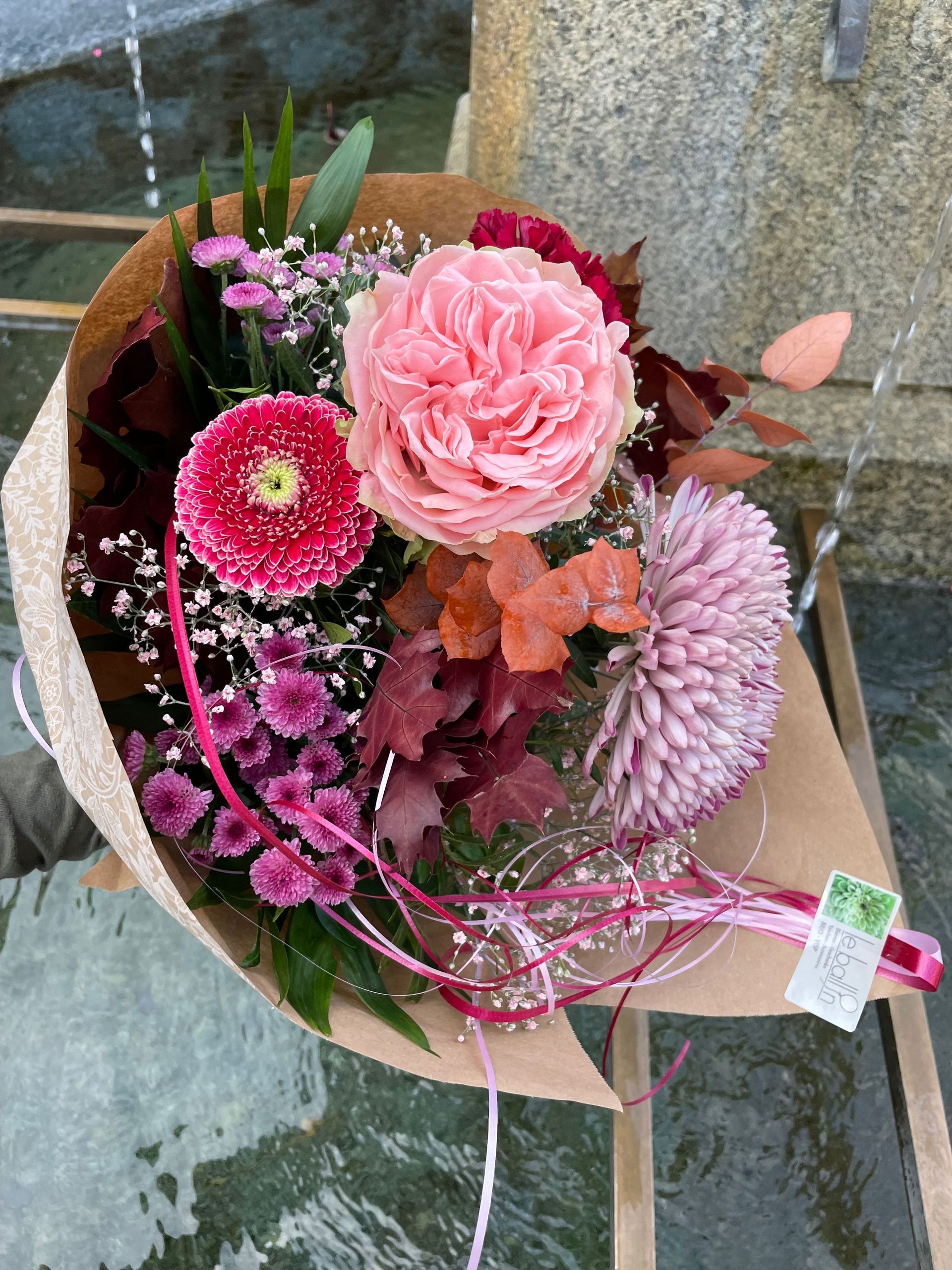 Auf einem Tisch steht ein Strauß rosa Blumen, in braunes Papier eingewickelt.