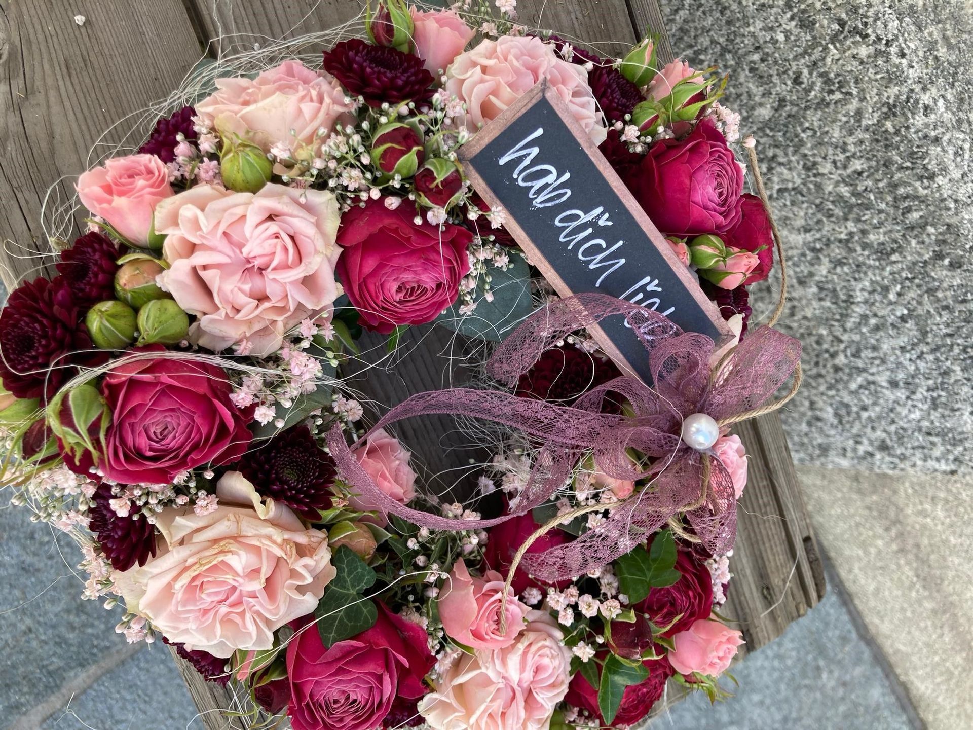 Ein Kranz aus rosa und weinroten Blumen mit einem Schild darauf.