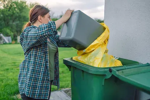 Una mujer está metiendo un bote de basura en un bote de basura verde.