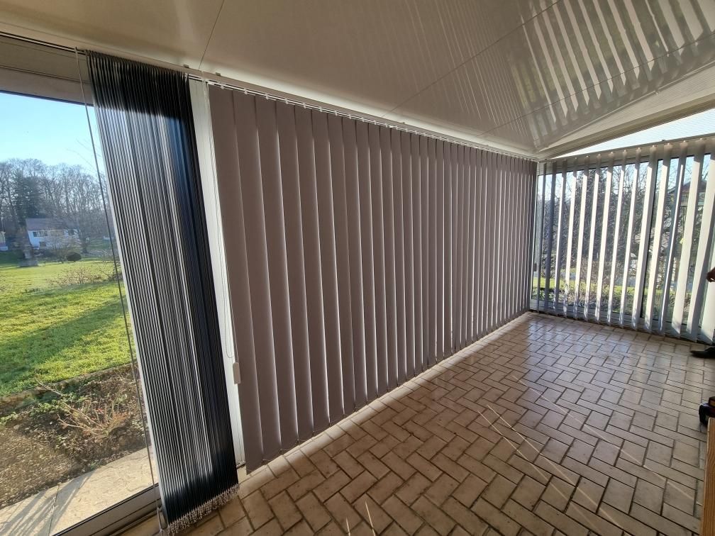 Terrasse couverte avec sol en briques, stores verticaux et vue sur un jardin.