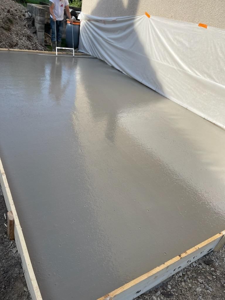 Dalle de béton fraîchement coulée en train de sécher contre un mur recouvert d'une bâche, avec un ouvrier à l'extrémité opposée.