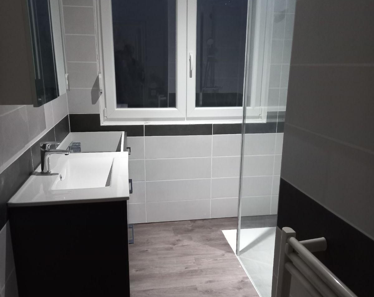 Salle de bains moderne avec lavabo noir et blanc, carrelage gris, fenêtre, paroi de douche en verre et sèche-serviettes.