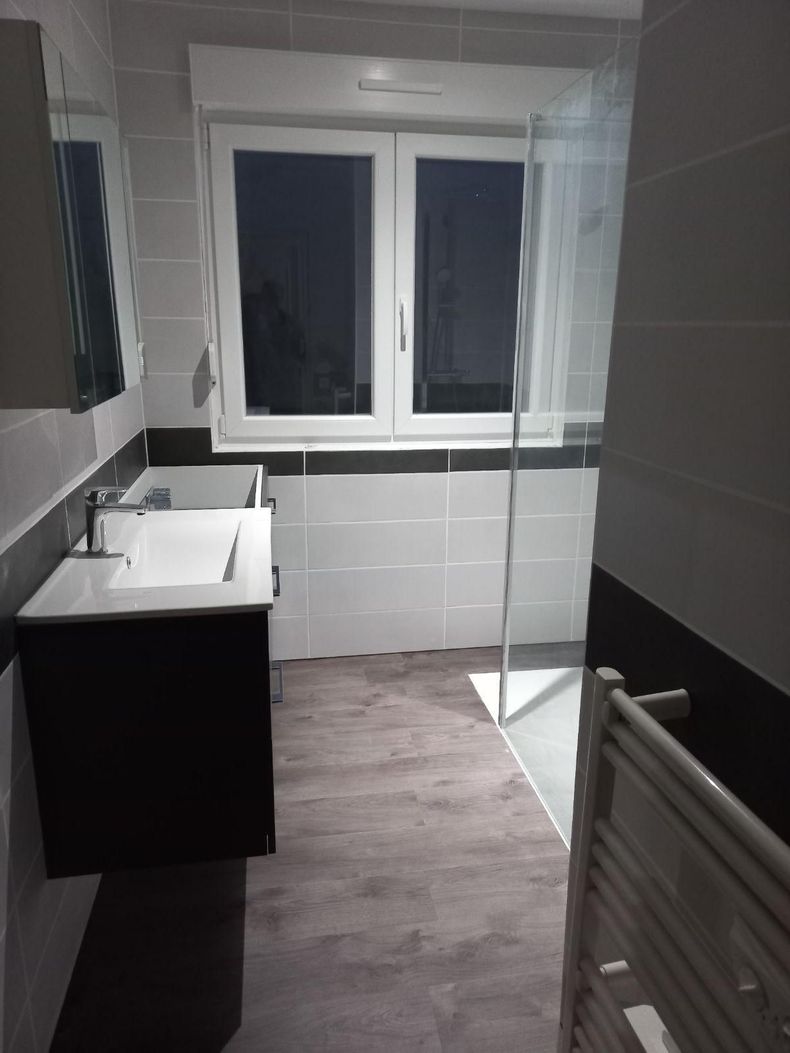 Salle de bains avec lavabo noir et blanc, carrelage gris, fenêtre, paroi de douche en verre et sèche-serviettes.