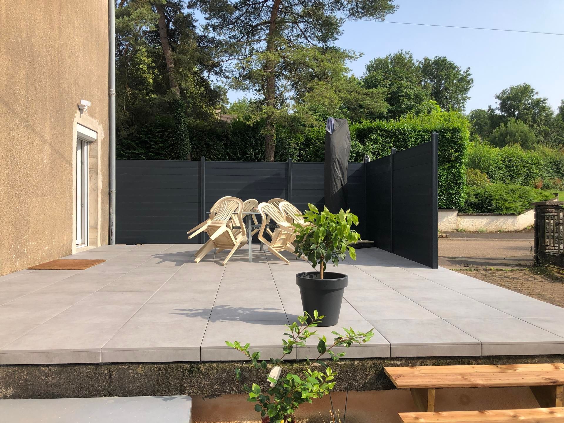 Terrasse extérieure avec carrelage gris, clôture noire, chaises blanches et plante en pot à côté d'une maison.