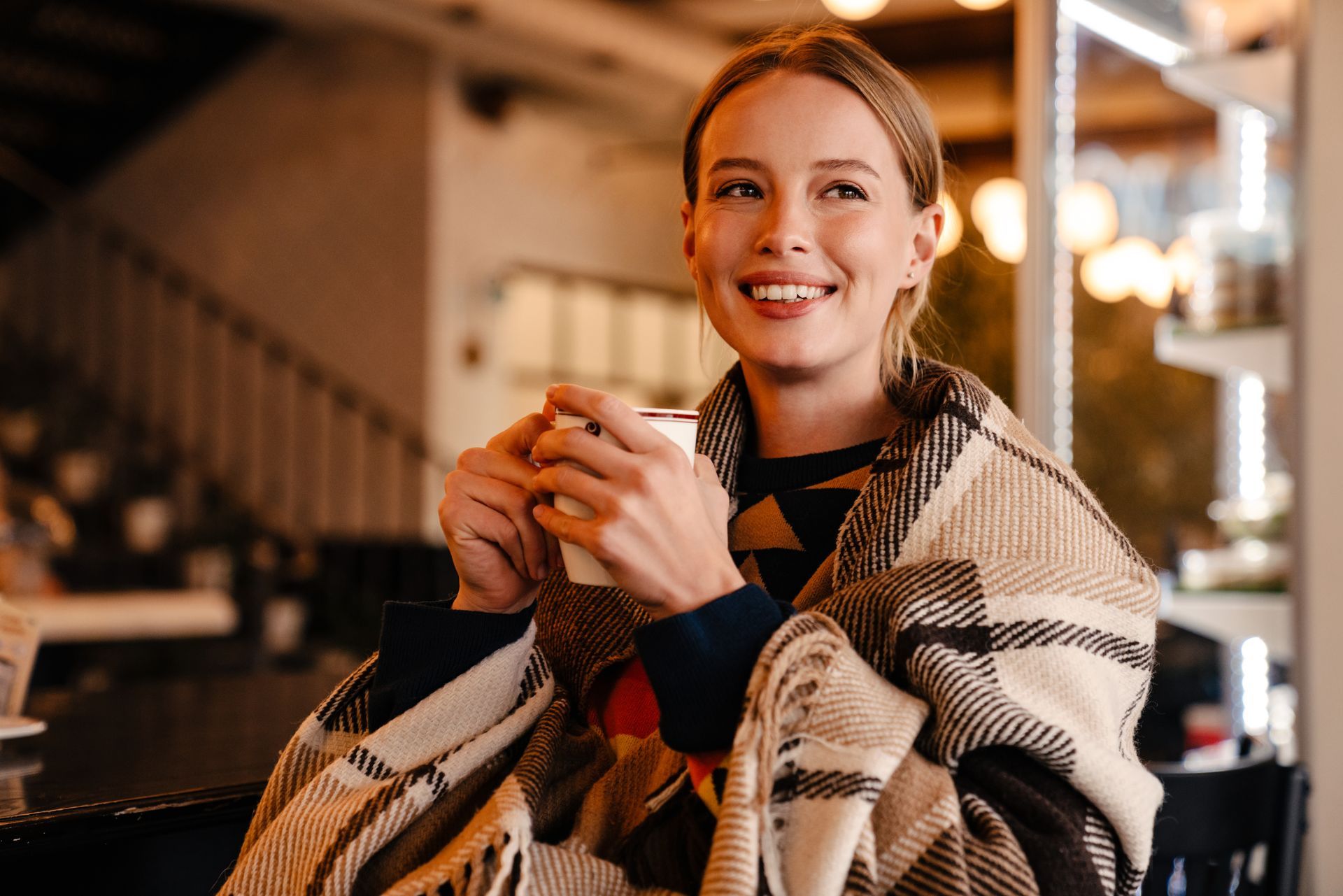 Une personne souriante, tenant une tasse dans un café, enveloppée dans un châle à carreaux.