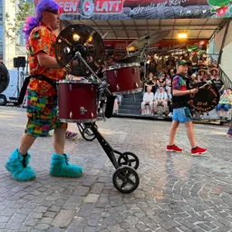 Ein Mann in einem Clownskostüm spielt auf einem Wagen Schlagzeug.