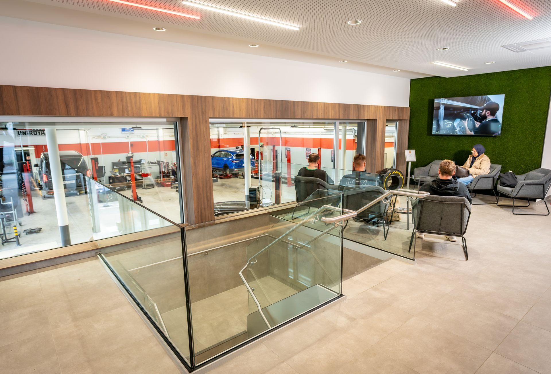 Modern office lobby with glass railings, lounge seating, and people sitting near a wall-mounted TV