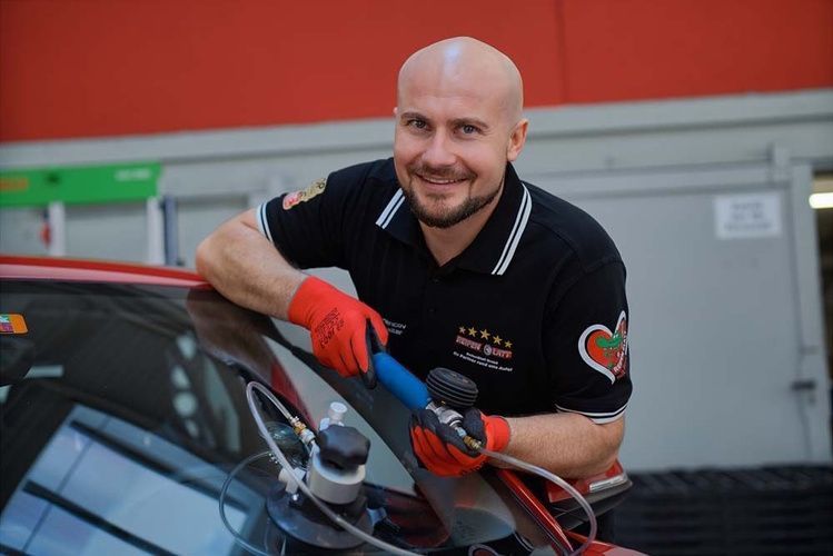 Ein lächelnder Mechaniker mit roten Handschuhen poliert in einer Garage eine Autoscheibe.