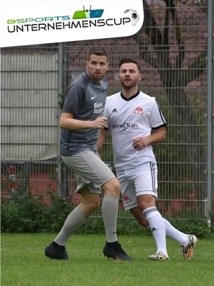 Zwei Männer spielen auf einem Feld vor einem Zaun Fußball.