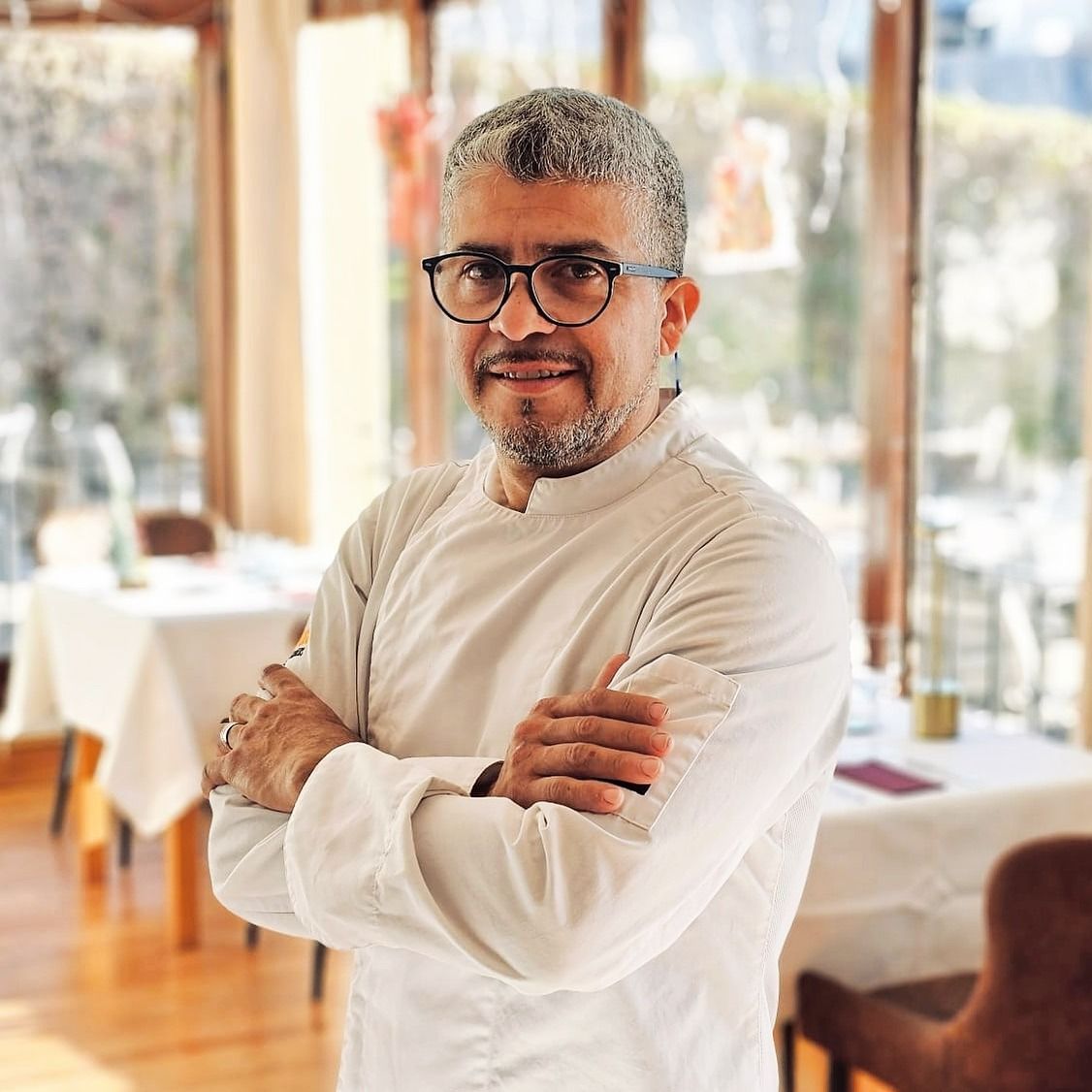 Un hombre con gafas y barba está de pie con los brazos cruzados en un restaurante.