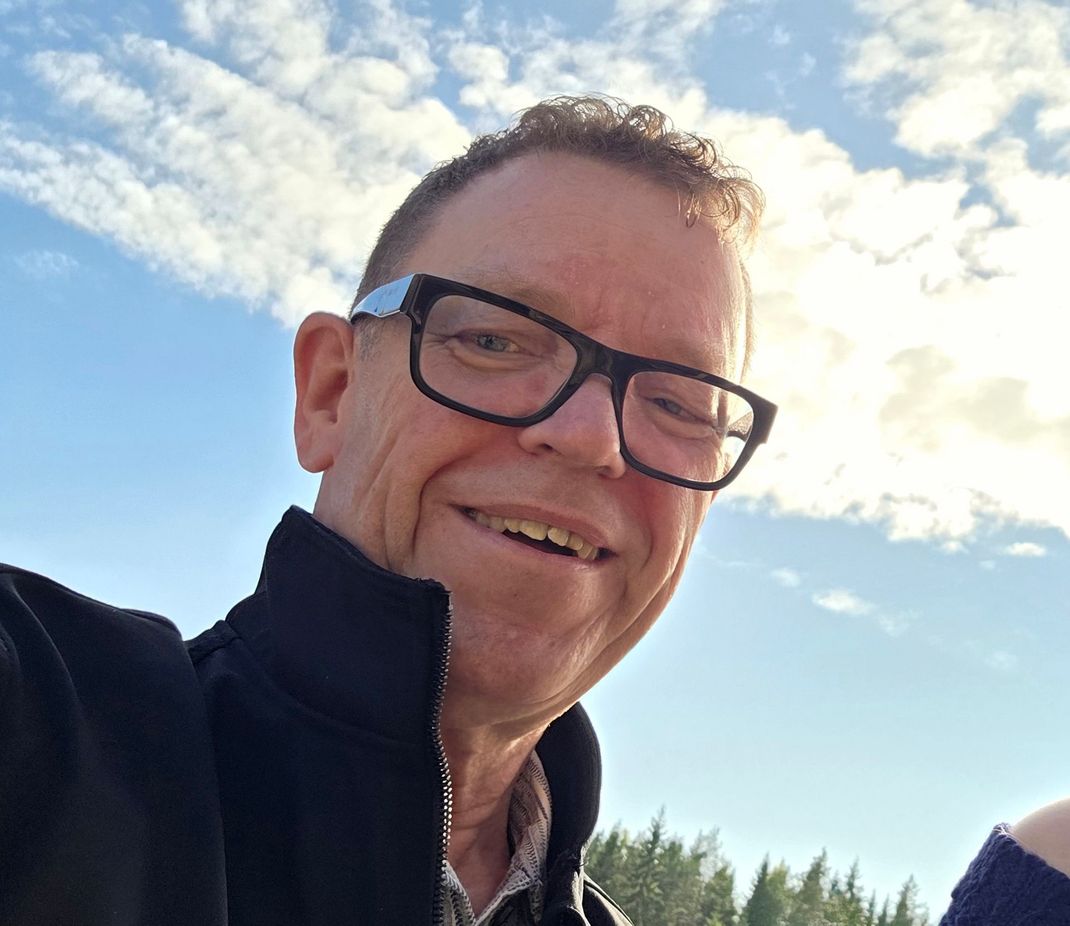 Tervetuloa kyytiin! Aurinkolasit päässä oleva mies hymyilee veneessä, jossa on vettä, veneitä ja kivinen rakennelma, jonka taustalla on torni.