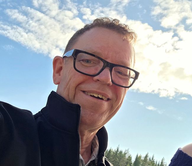 Tervetuloa kyytiin! Aurinkolasit päässä oleva mies hymyilee veneessä, jossa on vettä, veneitä ja kivinen rakennelma, jonka taustalla on torni.