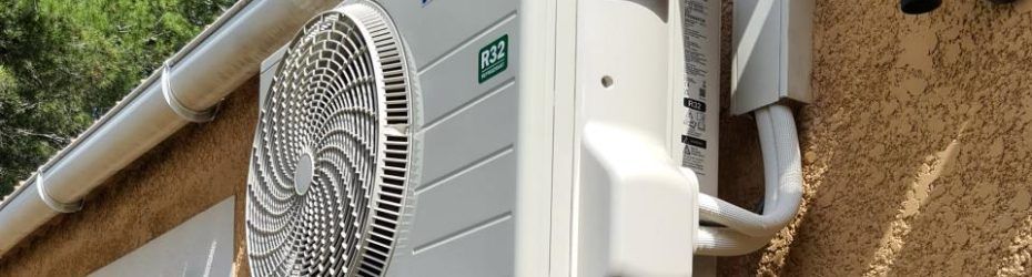 Unité de climatisation extérieure fixée sur un mur beige avec tuyauterie blanche apparente.