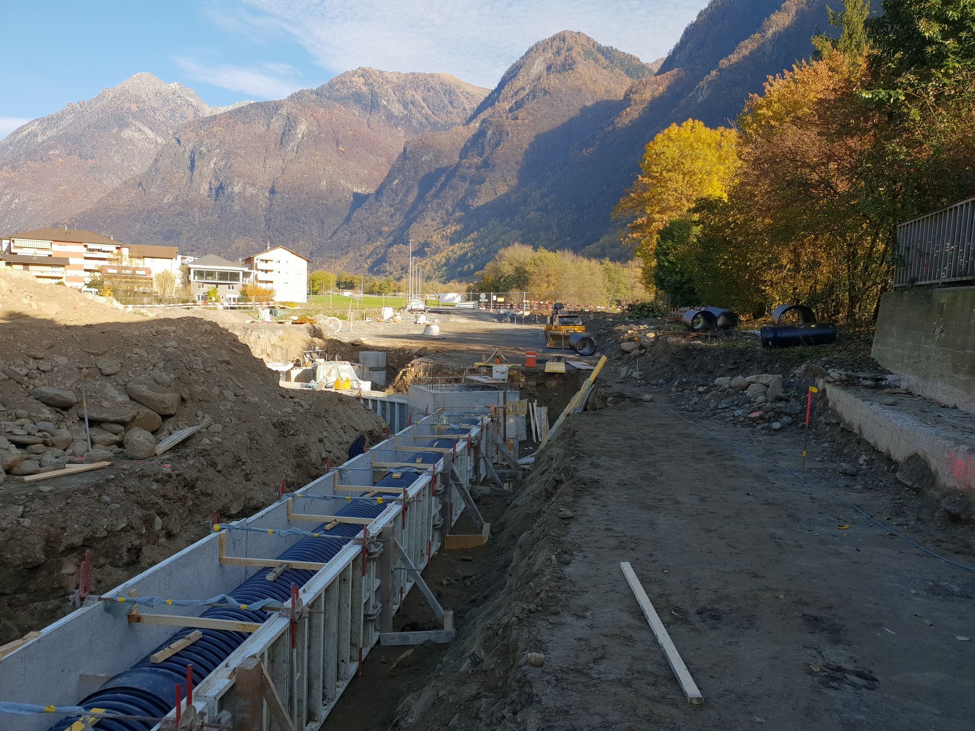 Un cantiere edile con una lunga trincea di fondazione in cemento armato in primo piano, sullo sfondo di un paesaggio montano.