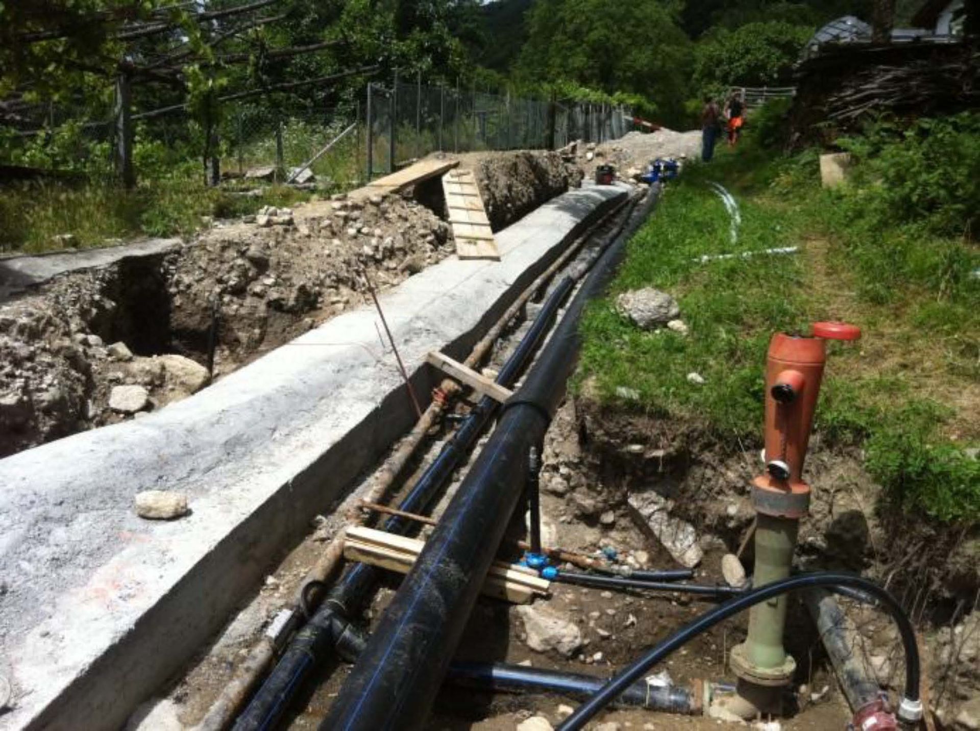 Lungo una trincea di cemento in costruzione, accanto a un pendio erboso e a un idrante rosso, si snodano tubi neri dell'acqua.