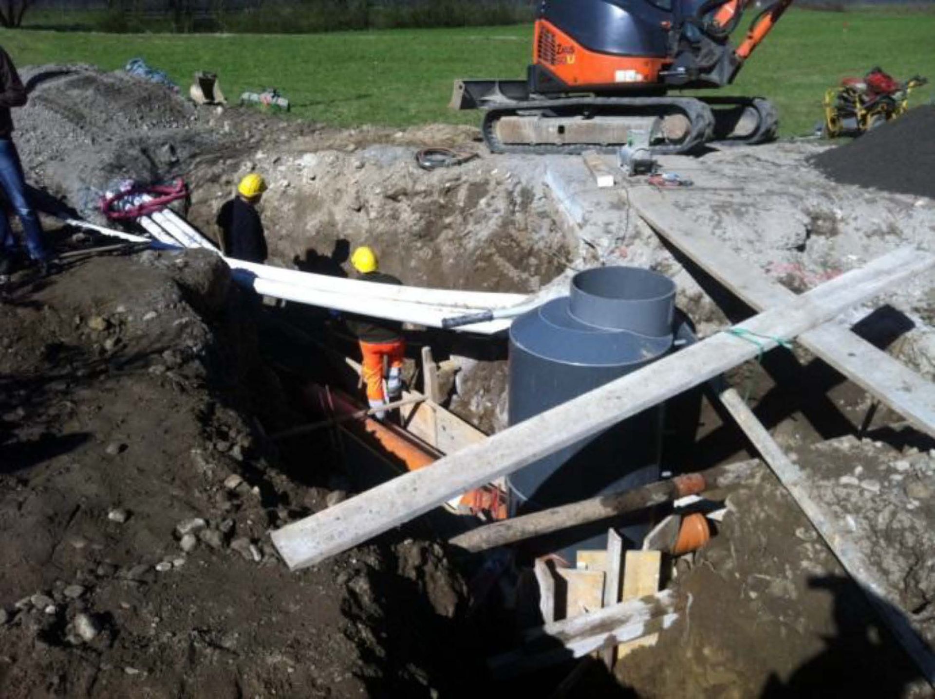 Operai edili in una trincea intenti a installare tubature vicino a un grande serbatoio cilindrico grigio, con un escavatore nelle vicinanze.