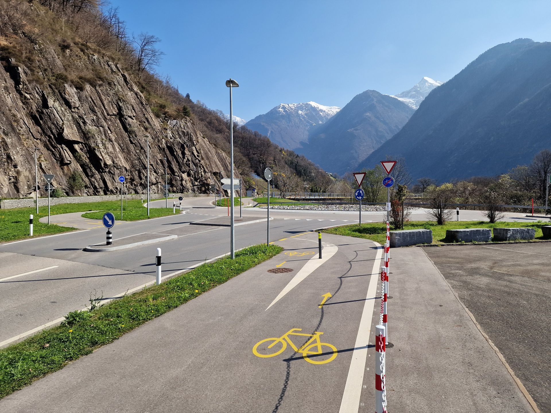 In una giornata di sole, una pista ciclabile asfaltata con segnaletica gialla conduce a un incrocio di montagna con cartelli stradali.