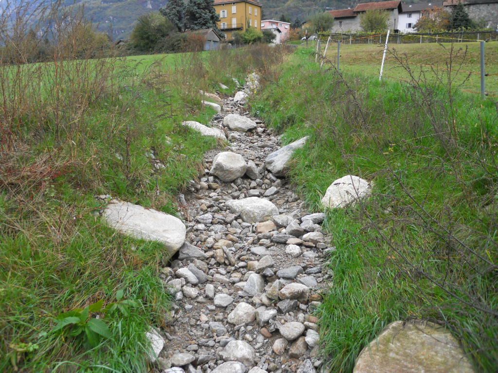 Un letto di torrente asciutto e roccioso che serpeggia attraverso un campo erboso, con case visibili sullo sfondo.