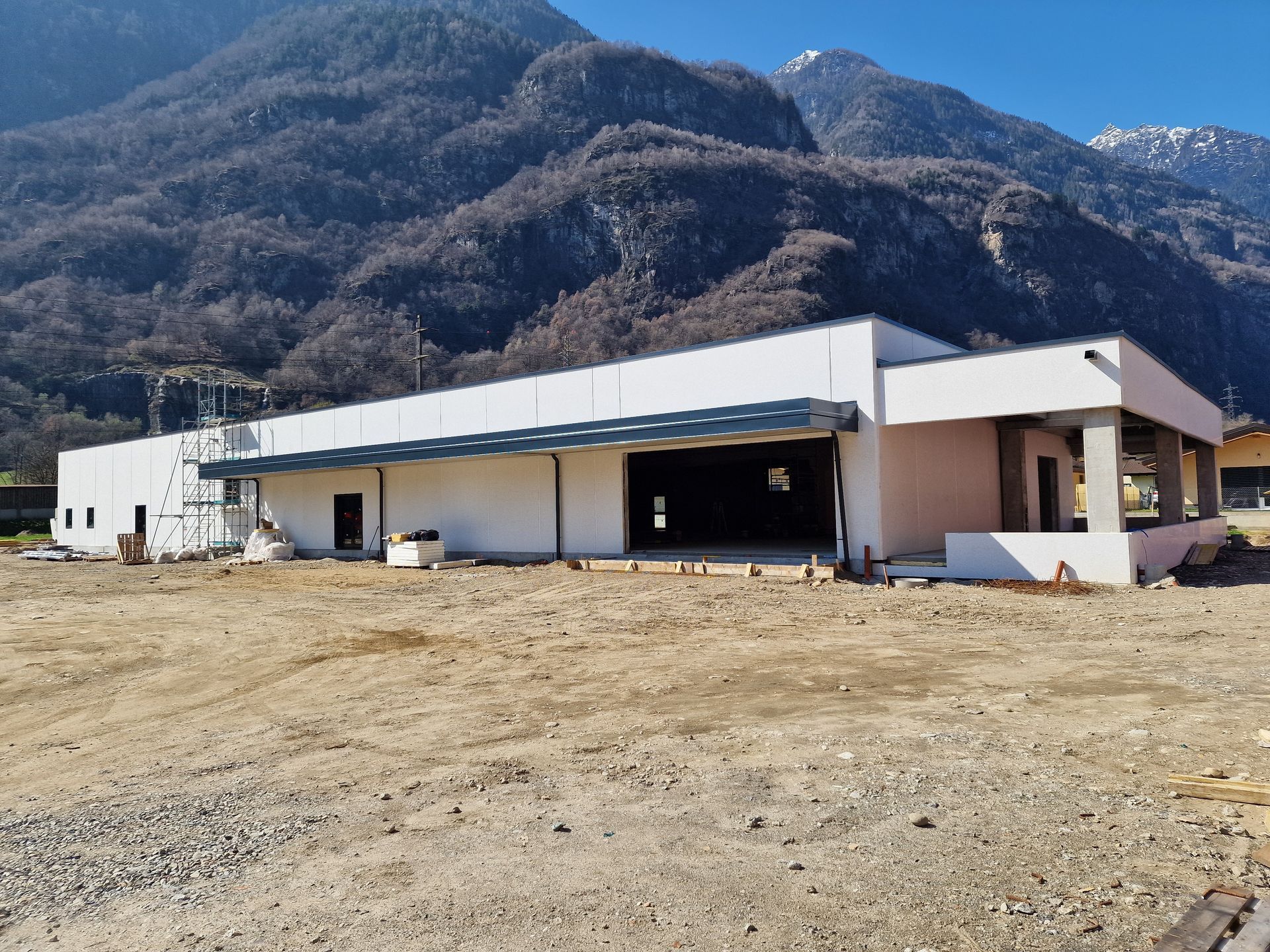 Un lungo edificio bianco e moderno, ancora in costruzione, sorge su un terreno sterrato di fronte a un'imponente e aspra montagna.