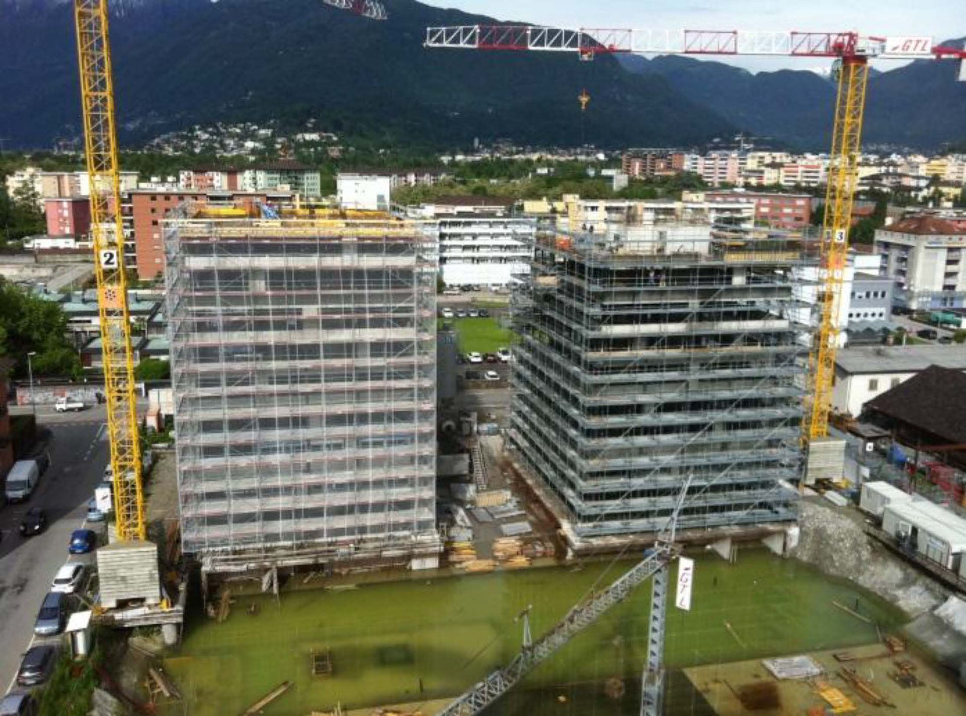Due alti edifici in cemento in costruzione, circondati da impalcature e gru gialle, con le montagne sullo sfondo.
