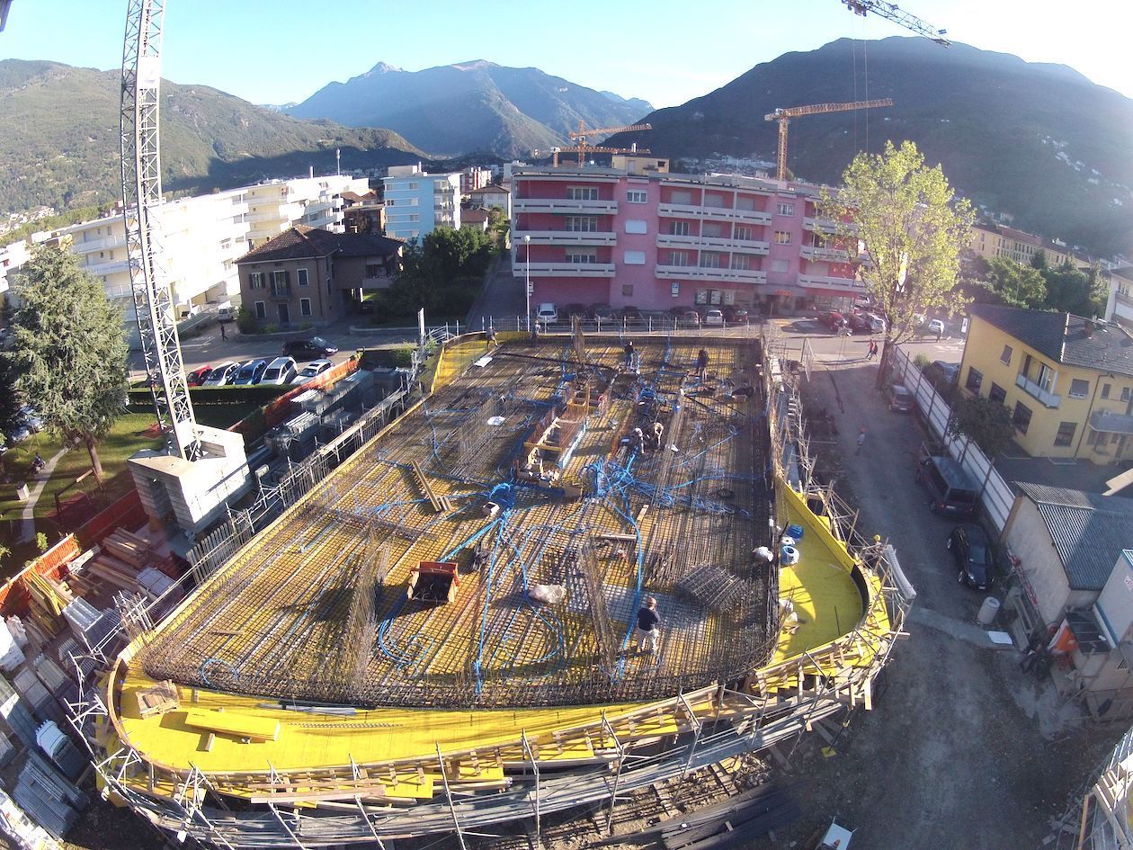 Veduta aerea di un cantiere edile in una zona montuosa, con fondamenta delimitate da un bordo giallo, barre d'acciaio per cemento armato e un'alta gru.