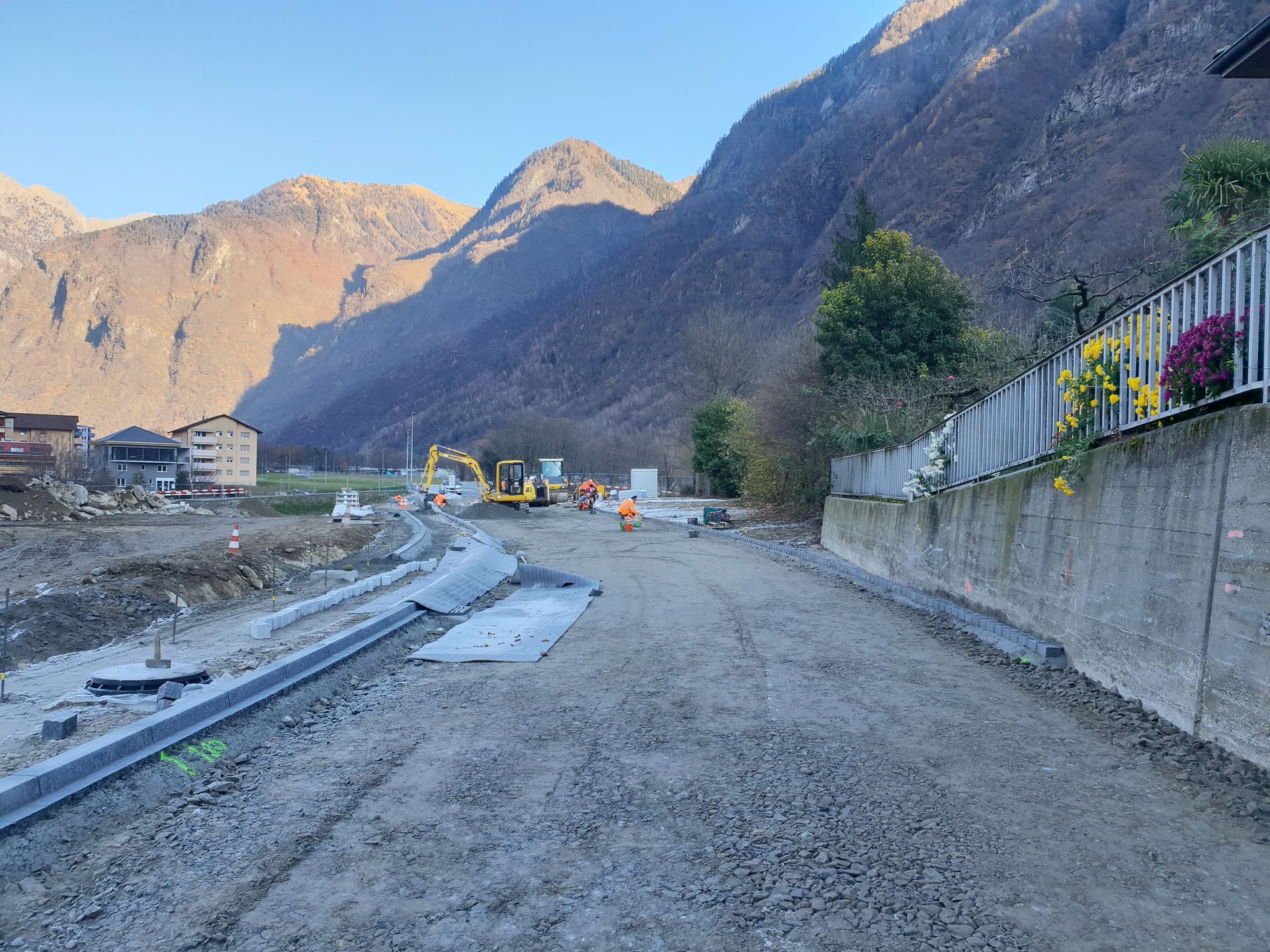 Cantiere edile con escavatore e terreno ghiaioso ai piedi delle montagne, sotto un cielo limpido.