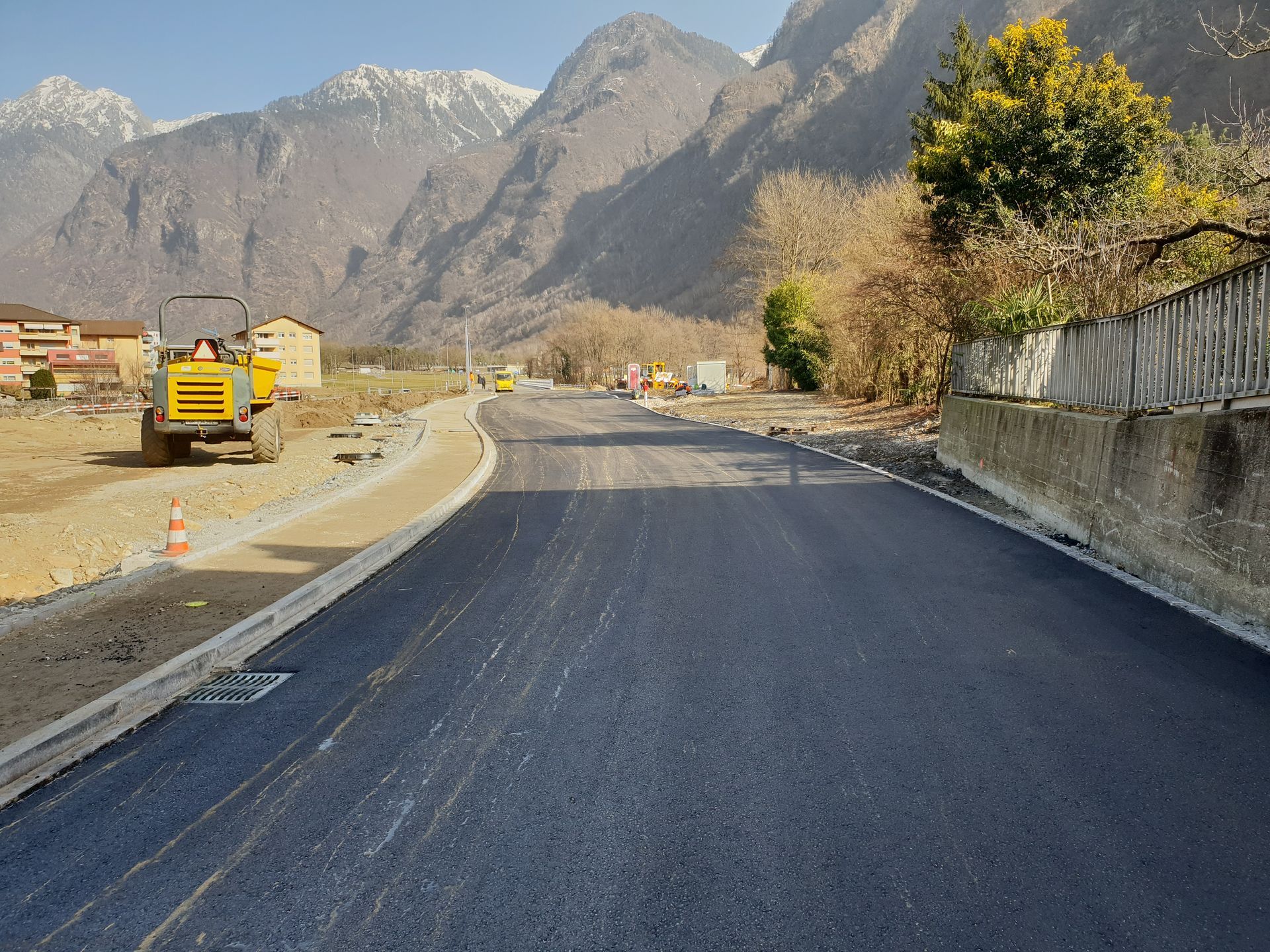 Una strada appena asfaltata conduce verso una catena montuosa, con un veicolo da cantiere giallo parcheggiato a lato.