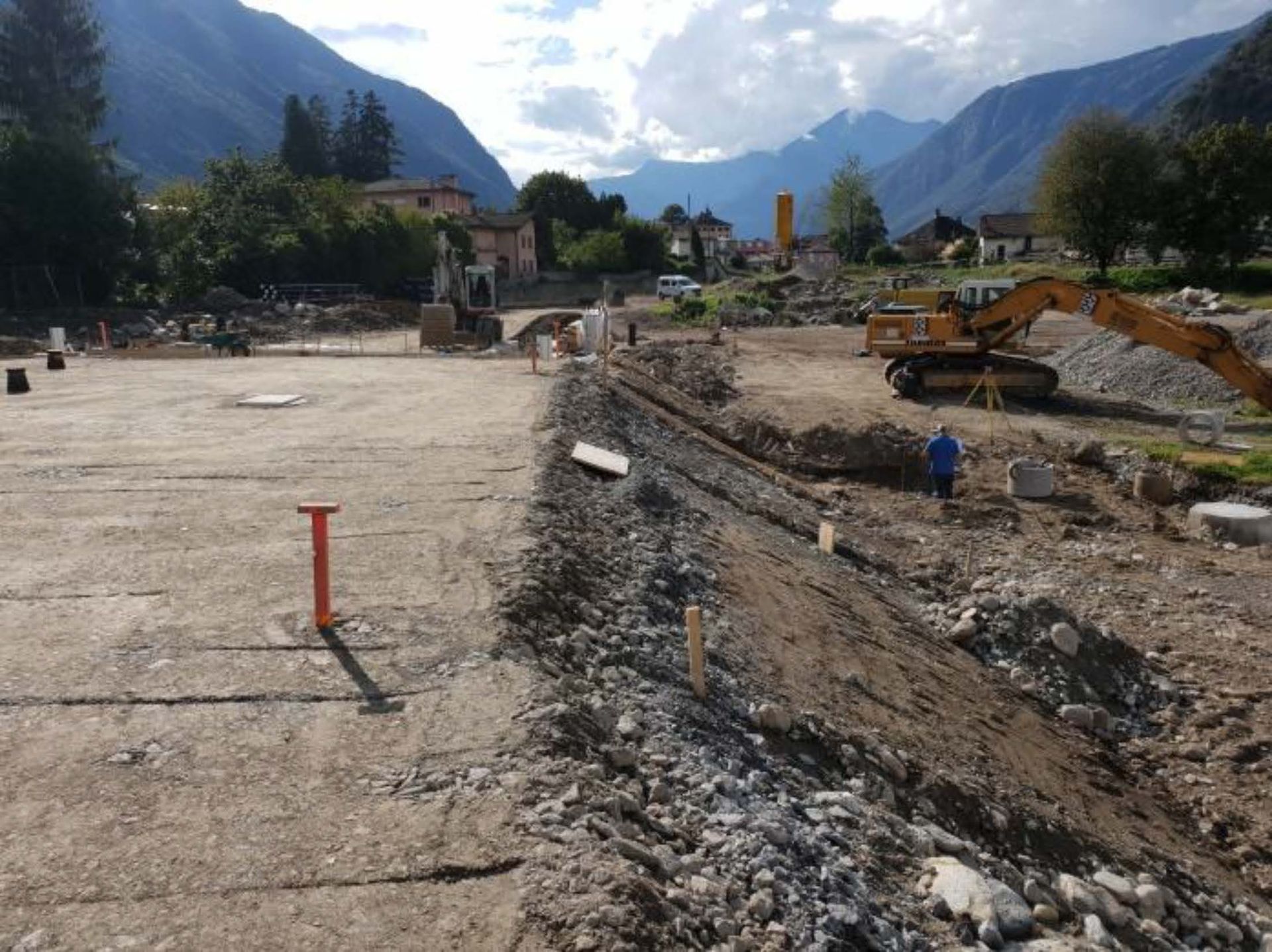 Cantiere edile con superficie pianeggiante, terrapieno in pendenza e macchinari pesanti, sullo sfondo di un paesaggio montuoso.