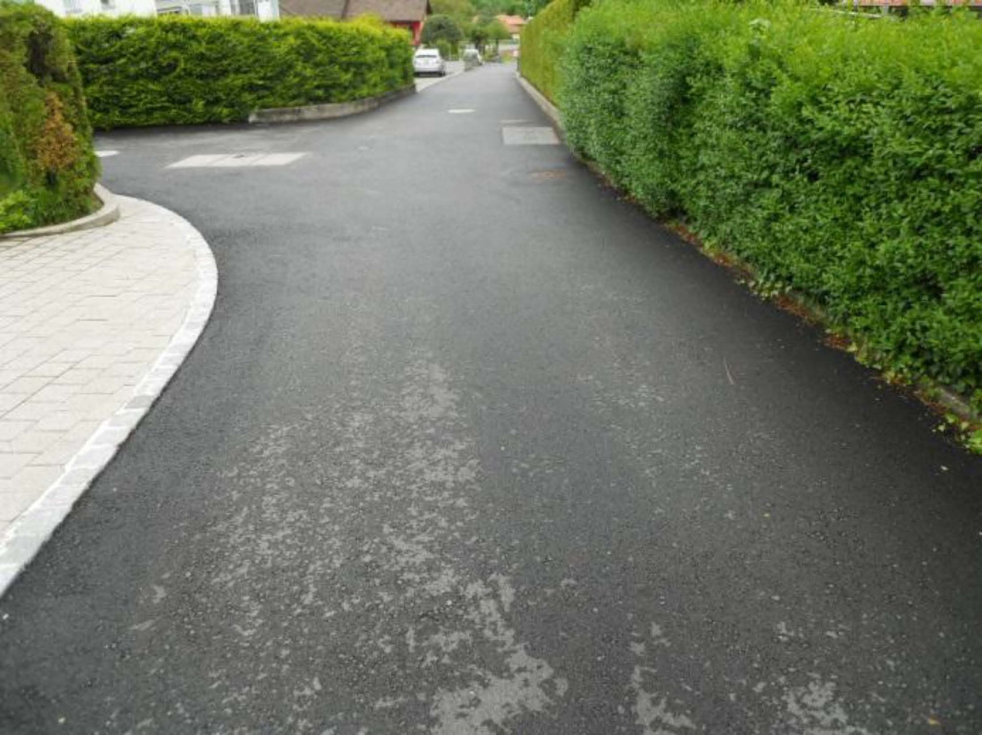 Un vialetto pavimentato curva costeggiando un sentiero pedonale lastricato sulla sinistra e una lunga siepe verde ben curata sulla destra.