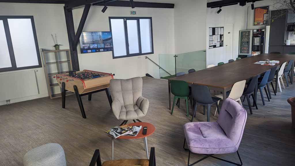 Un espace de détente lumineux et ouvert, avec un baby-foot, une grande table de conférence en bois et des chaises colorées sur un parquet.