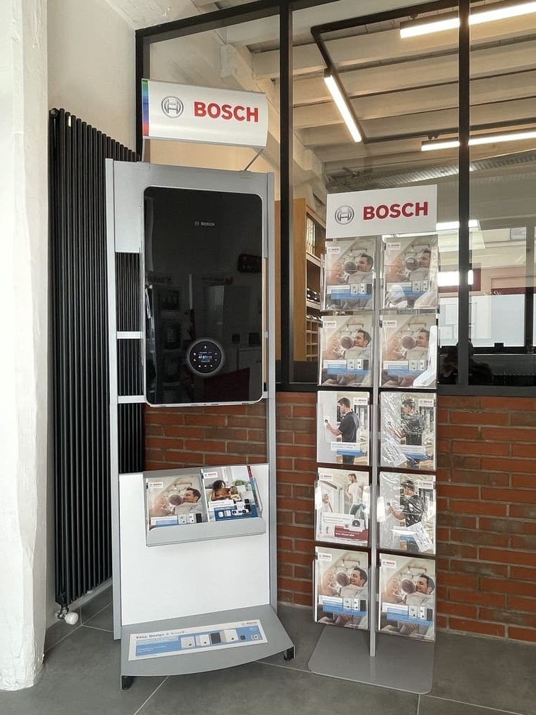 Un présentoir promotionnel Bosch mettant en scène une chaudière murale noire et un présentoir de brochures dans un magasin aux murs de briques.