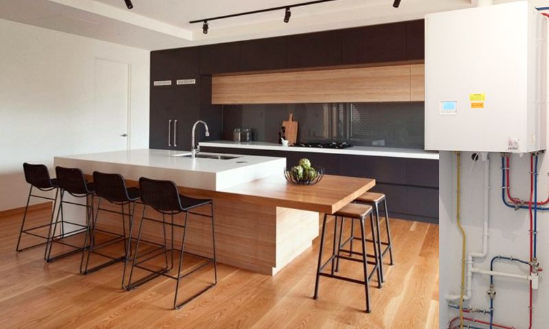 Un îlot de cuisine moderne avec tabourets de bar précède des armoires sombres et un meuble utilitaire mural blanc avec tuyauterie.