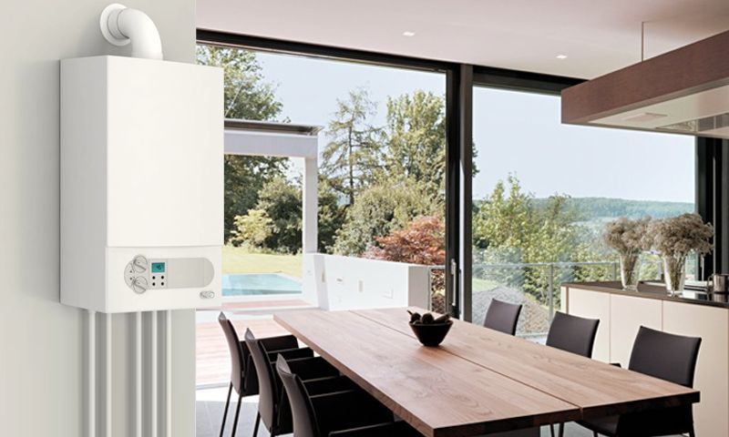Une chaudière blanche murale installée dans une salle à manger moderne avec une table en bois et de grandes fenêtres donnant sur un jardin.