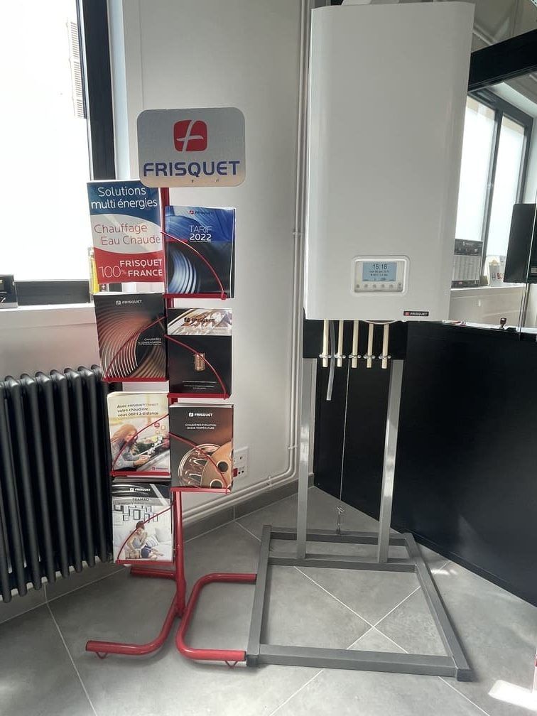 Dans un bureau, une chaudière Frisquet blanche fixée au mur repose sur un support métallique, à côté d'un présentoir à brochures rouge.