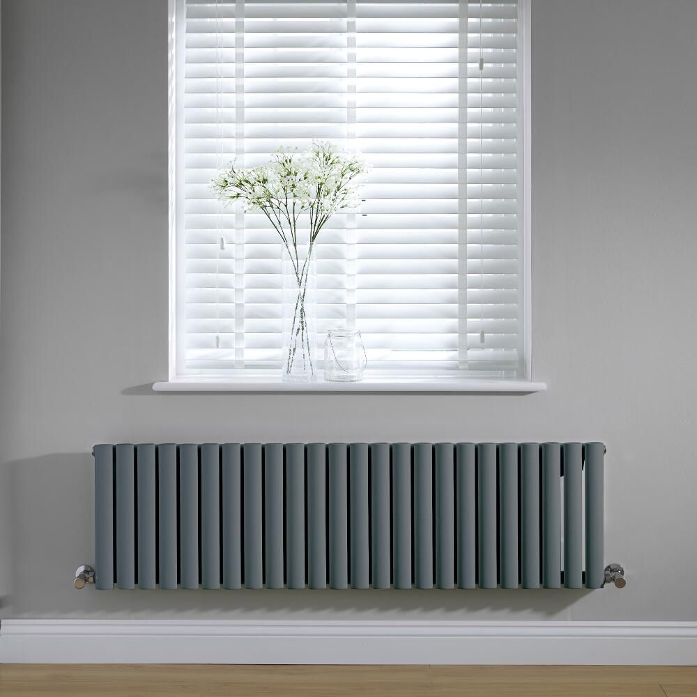 Un radiateur horizontal gris foncé, fixé sur un mur gris sous une fenêtre avec des stores blancs et un vase de fleurs blanches.