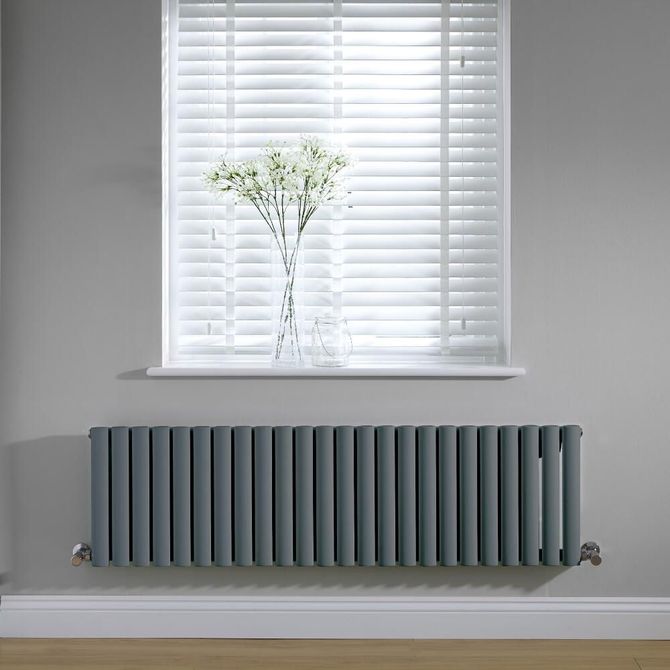 Un radiateur horizontal gris foncé, fixé sur un mur gris sous une fenêtre à stores blancs et un vase de fleurs.