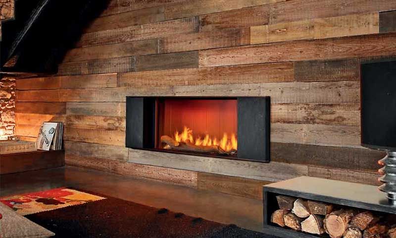 Une cheminée moderne encastrée dans un mur rustique en planches de bois, avec un support métallique pour le bois de chauffage à proximité.