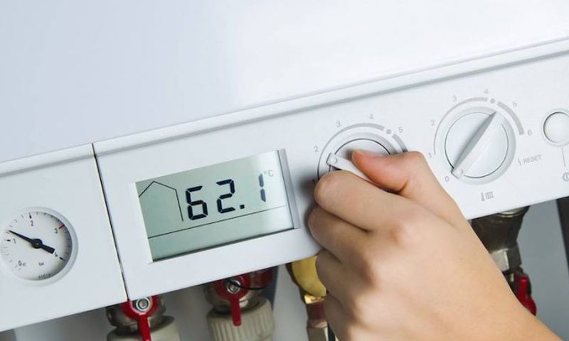 Une personne ajuste les paramètres d'une chaudière à gaz murale blanche dont l'affichage numérique indique 62,1 degrés.