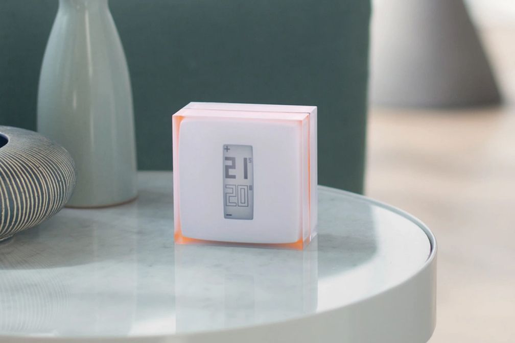 Une horloge numérique carrée blanche, ornée d'un liseré orange, est posée sur une table ronde blanche à côté d'un vase décoratif.