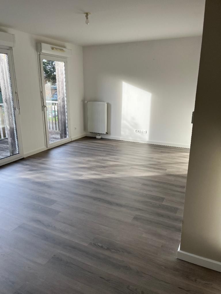 Chambre vide avec sol stratifié gris, murs blancs et radiateur. La lumière du soleil entre par un balcon.