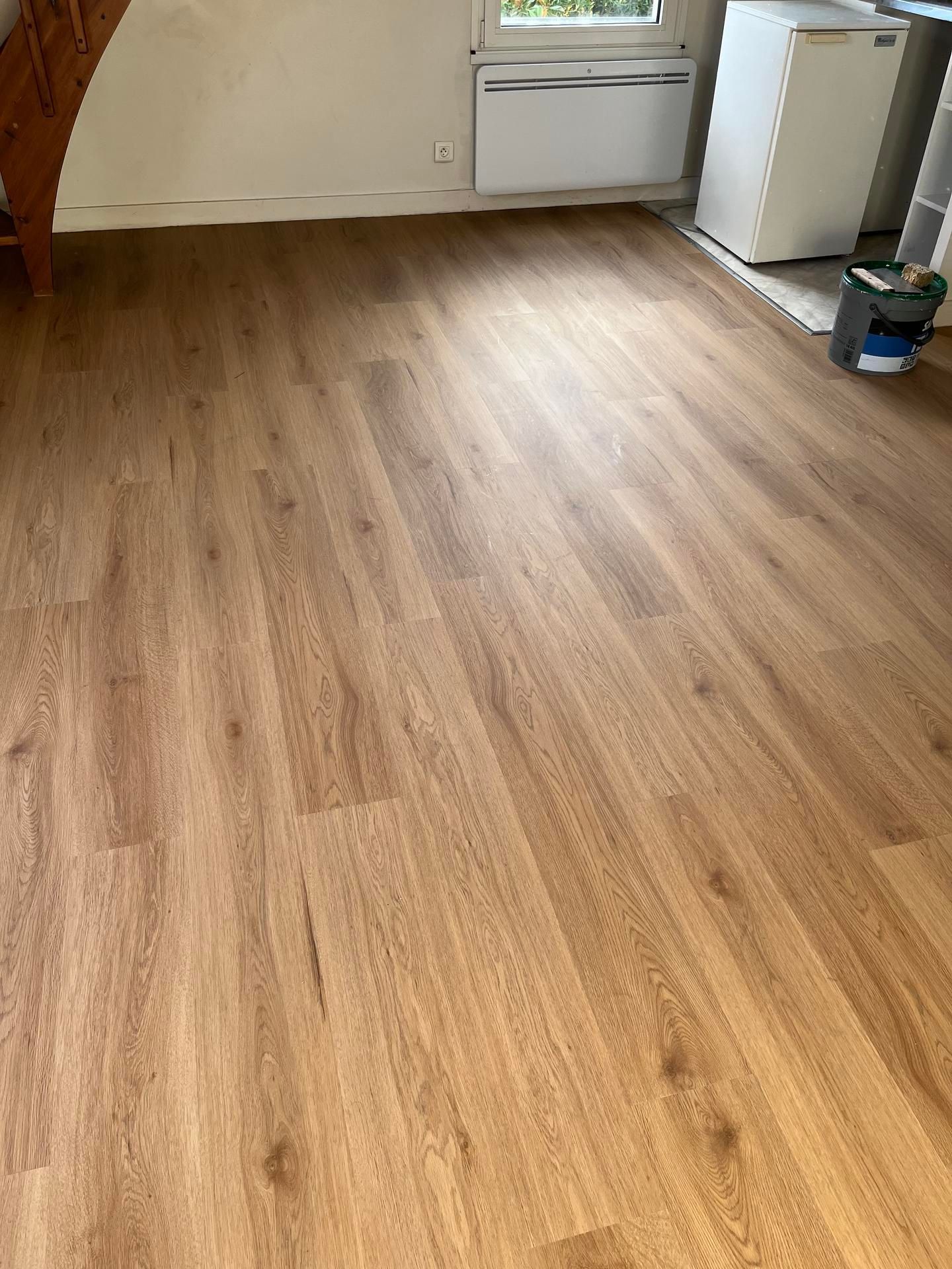 Parquet aspect bois dans une pièce avec un réfrigérateur blanc et une fenêtre visibles.