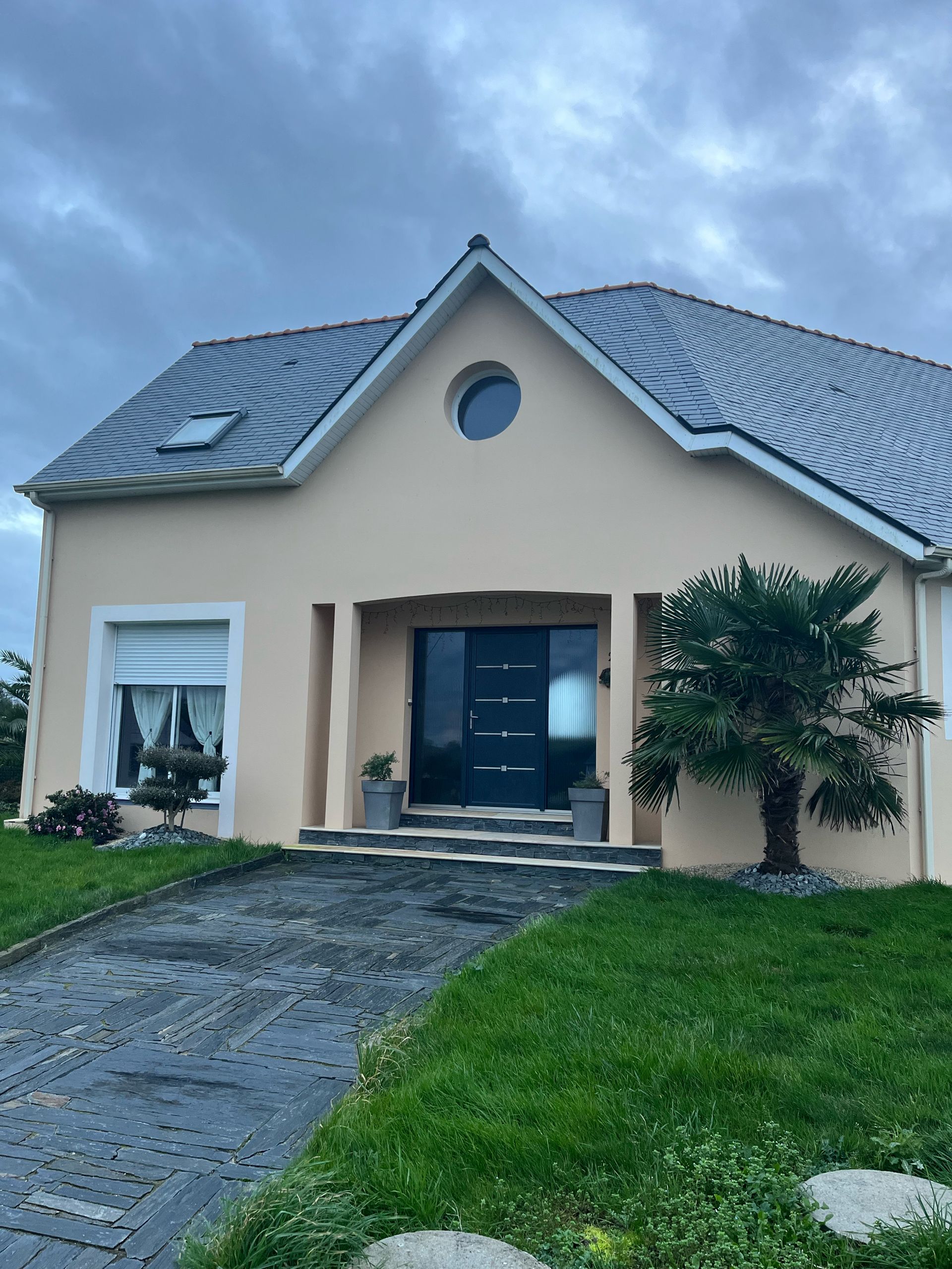 Maison en stuc beige avec porte sombre, toit gris et une fenêtre circulaire au-dessus de l'entrée.