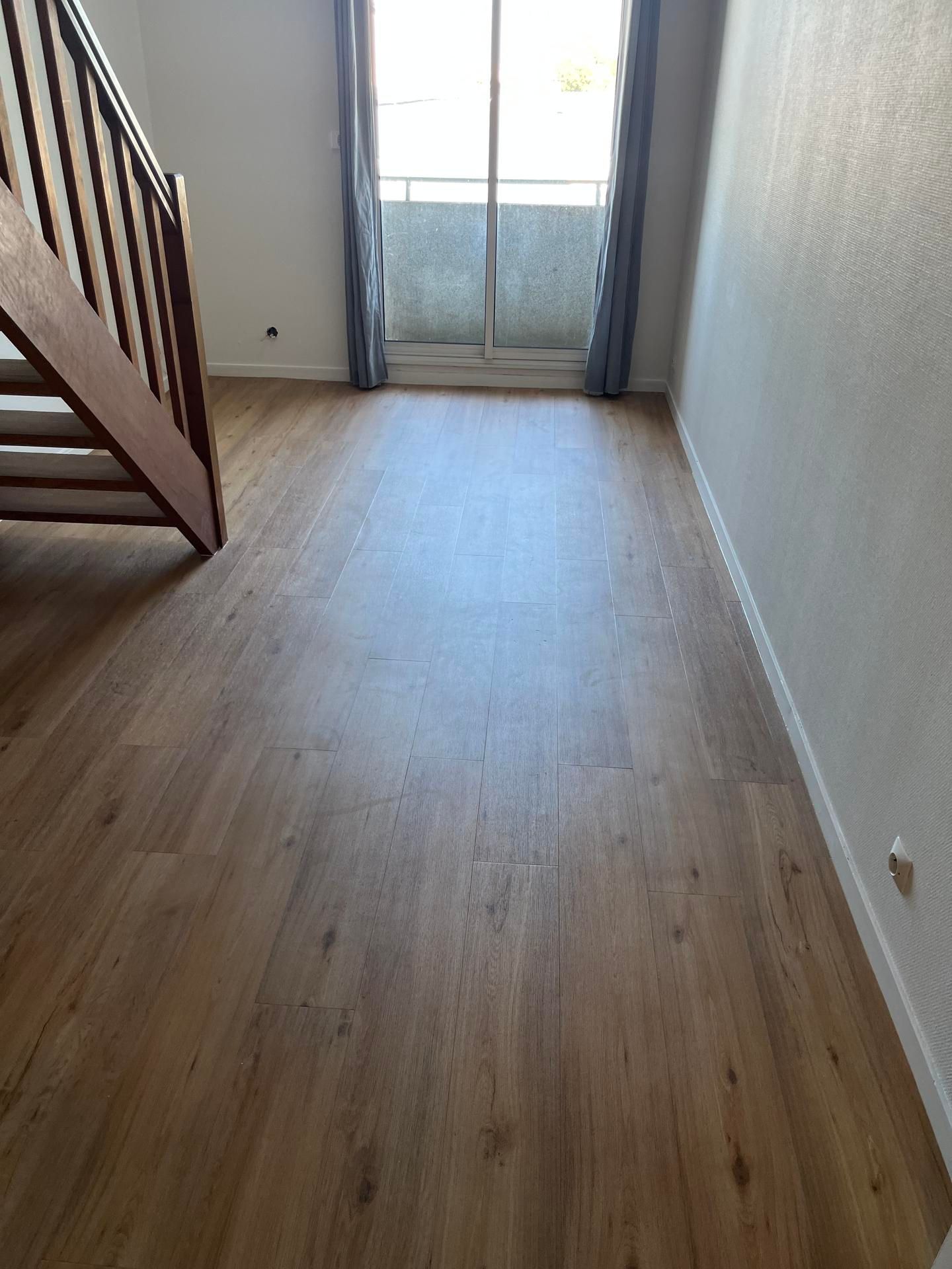 Pièce vide avec parquet imitation bois, escalier à gauche, fenêtre avec rideaux transparents à l'arrière.