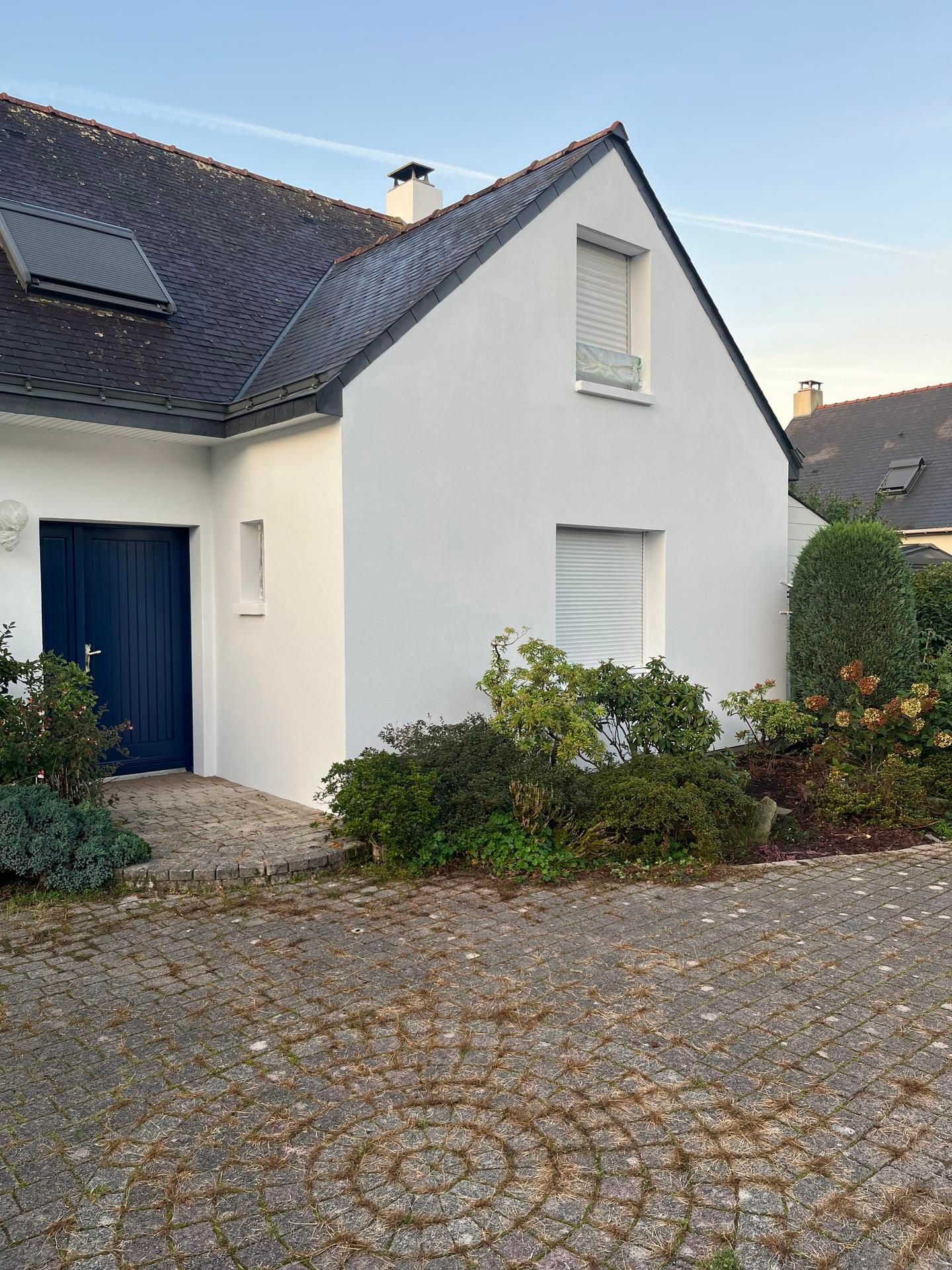 Maison blanche avec une porte bleu foncé et deux fenêtres, entourée d'aménagement paysager, d'une allée pavée et d'un toit gris.