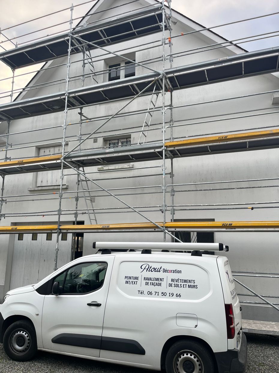 Échafaudage sur un bâtiment blanc en rénovation. Buissons et gravier au premier plan. Ciel couvert.