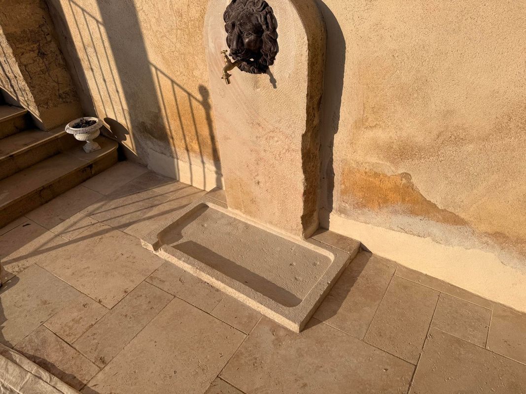 Fontaine en pierre à tête de lion, encastrée dans un mur beige patiné. Un bassin se trouve à sa base.