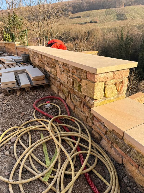 Mur de soutènement en pierre avec des dalles de couverture claires. Des tuyaux d'arrosage, des palettes et un flanc de colline sont visibles en arrière-plan.