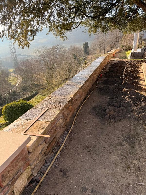 Un long mur de soutènement en pierre patinée par le temps, offrant une vue panoramique ; des arbres dénudés et un paysage lointain.