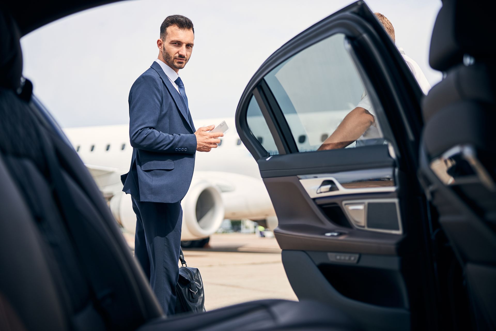 Photo dun homme en costume qui s'apprète à monter dans une voiture. Avion au sol en arrière plan.