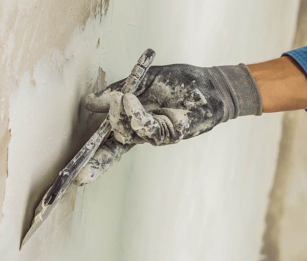 Eine behandschuhte Hand benutzt einen Spachtel, um Spachtelmasse an eine Wand aufzutragen.